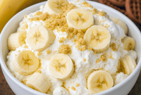 Glass bowl of banana pudding fluff layered with vanilla wafers, sliced bananas, and mini marshmallows, topped with a whole wafer.