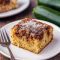 Slice of cinnamon zucchini coffee cake with a golden crumb topping on a plate, steam rising and a coffee cup in the background.