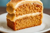 Slice of applesauce spice cake topped with cinnamon cream cheese frosting and chopped pecans on a ceramic plate.