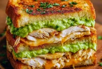 Toasted sandwich filled with sliced grilled chicken, ripe avocado, melted cheese and tomato, halved and stacked on a wooden board.