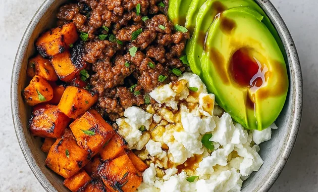 Overhead shot of a bowl with seasoned ground beef, roasted sweet potato cubes, sliced avocado, dollops of cottage cheese, and a honey drizzle, garnished with cilantro.