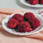Close-up of glossy red velvet cheesecake bites coated in fine red cake crumbs, arranged on a white serving platter with a drizzle of white chocolate.