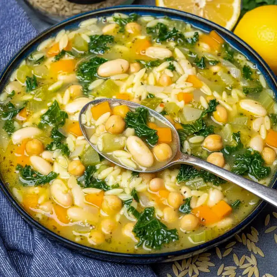 Steaming bowl of orzo vegetable soup with chickpeas, kale, lemon wedge, and a slice of crusty bread on a wooden board, Vegan Soup Recipes.