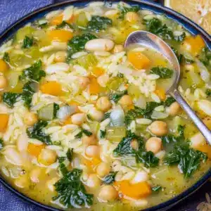 Steaming bowl of orzo vegetable soup with chickpeas, kale, lemon wedge, and a slice of crusty bread on a wooden board.