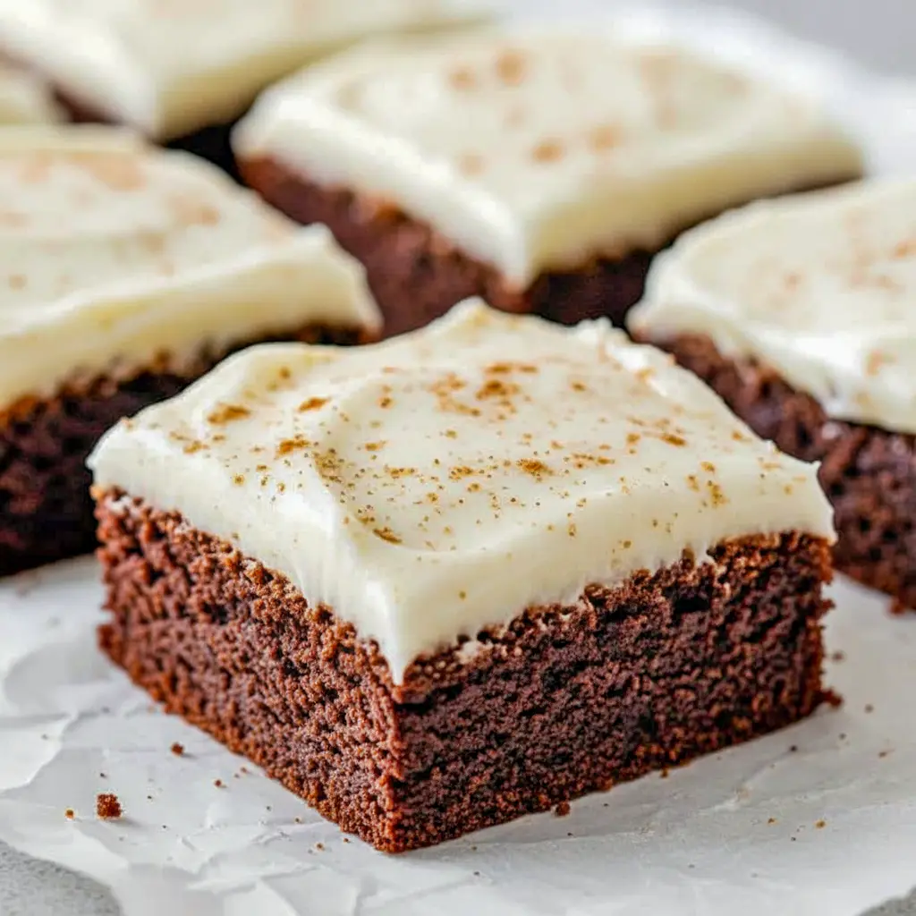 A platter of square gingerbread brownies frosted with creamy eggnog cream-cheese icing, dusted with cinnamon and arranged on a festive holiday napkin.