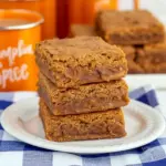 Stack of chewy pumpkin blondies with visible cinnamon chips, drizzled caramel, and a sprinkle of flaky sea salt on a rustic wooden board.