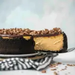 Slice of espresso cheesecake on a white plate with Oreo crust, topped with chocolate ganache and chocolate shavings.