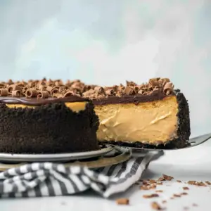 Slice of espresso cheesecake on a white plate with Oreo crust, topped with chocolate ganache and chocolate shavings.
