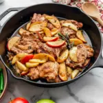 Golden skillet with seared chicken thighs, sautéed apple slices and shallots in a glossy apple-cider sauce, garnished with thyme.