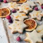 Overhead shot of star-shaped cranberry orange shortbread cookies glossed with orange glaze and sprinkled with zest, arranged on a wooden board with a festive ribbon nearby, Orange Christmas Recipes.