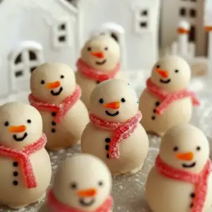 Close-up of three dairy-free white chocolate snowman truffles with tiny candy scarves and piped faces on a festive plate, No Bake Desserts For Christmas.