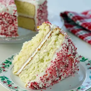 Slice of peppermint layer cake with white buttercream, crushed candy-cane rim, and a sprig of evergreen on a festive plate, Peppermint Cake.