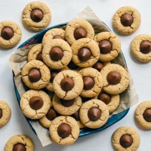 Golden peanut butter cookies rolled in sugar, each topped with a glossy milk chocolate kiss and stacked on parchment, Easy Peanut Butter Blossoms.