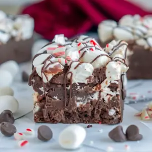 Close-up of peppermint chocolate fudge squares topped with mini marshmallows, crushed candy cane, and a chocolate drizzle, arranged on a festive napkin, Peppermint Chocolate Fudge.