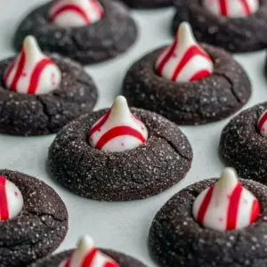 Close-up of a fudgy cookie with a white-striped Kiss pressed into the warm center, showcasing Chocolate Mint Blossoms with crushed candy cane, seasonal Peppermint Blossoms sparkle, and a single Peppermint Blossom in front, Chocolate Peppermint Blossoms.