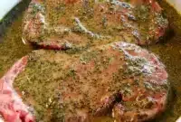Jar of Steak Marinade beside raw steaks on a cutting board, with fresh herbs, lemon, and a small bowl of sauce ready for brushing on the grill.