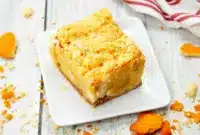 Close-up of a gooey Banana Blondies Recipe square on parchment, showing a golden vanilla-wafer crust, white chocolate pockets, and a fork ready to dig in.