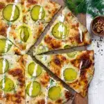 Top-down shot of a golden-brown pizza with garlic-white sauce, melted mozzarella, evenly spaced dill pickle slices, a drizzle of creamy garlic-dill sauce, and a sprinkle of fresh dill on a wooden pizza board, Dill Pickle Pizza Recipe.