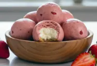 Close-up of Strawberry Cheesecake Protein Balls on parchment, pale pink centers with graham-cracker coating and a sprinkle of freeze-dried strawberry.
