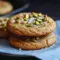 Golden Honey Pistachio Cookies dotted with chopped pistachios and a sprinkle of flaky sea salt on parchment paper.