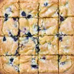 Plate of fudgy White Chocolate Blondies dotted with fresh blueberries and white chocolate chunks, cut into squares and stacked on parchment.