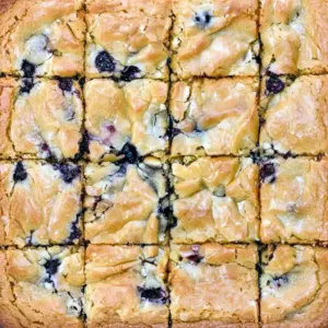 Plate of fudgy White Chocolate Blondies dotted with fresh blueberries and white chocolate chunks, cut into squares and stacked on parchment.