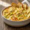 Bubbling Creamy Banana Pepper Chicken Dip in a small baking dish, melted golden cheese with visible banana pepper bits and a bowl of chips on the side — perfect Game Day Food and a top Party Food Appetizers choice.