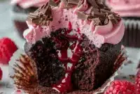 Close-up of Chocolate Lava Cupcakes split open to show a molten raspberry center, piped raspberry buttercream, fresh raspberry, and dark chocolate shavings on top.