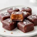 Close-up of dark chocolate–coated date caramels on parchment paper with one piece sliced to show the chewy caramel center and a sprinkle of flaky sea salt, Date Treats.