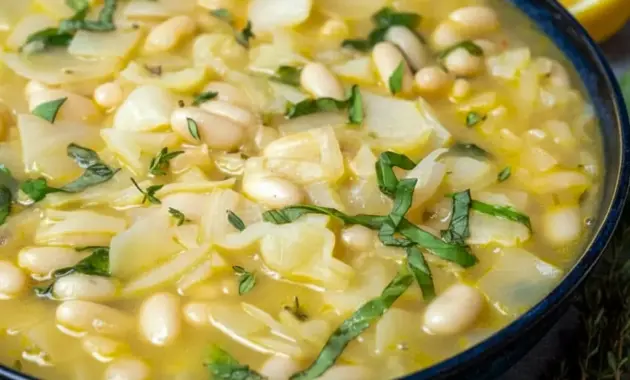 Bowl of steaming Bean And Cabbage Soup with shredded cabbage, white beans, fresh basil, and a lemon wedge, served with crusty bread.