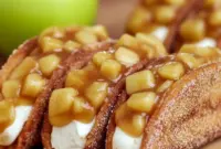 Homemade Apple Cheesecake Tacos: close-up of Cinnamon Sugar Tortillas shaped into taco shells, filled with creamy cheesecake and spiced apple topping, dusted with powdered sugar.