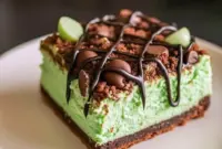 Slice of Mint Chocolate Cheesecake bar on a plate — vivid green mint cheesecake layer over a chocolate crumb crust, drizzled chocolate, and festive sprinkles for Saint Patrick's Day, a tempting example of elegant Cheesecake Bars.