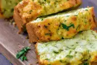 Slice of Zucchini Cheddar Bread on a wooden board, showing gooey cheddar and flecks of green zucchini.