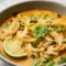 Steaming bowl of Thai Chicken Soup with coconut broth, rice noodles, shredded chicken, fresh basil, cilantro, scallion, and a lime wedge — top-down close-up.