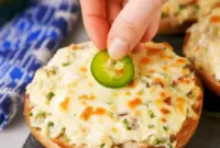 Close-up of baked Jalapeño Popper Bagels — bagel halves spread with jalapeño cream cheese, melted Monterey Jack, and bacon crumbles, garnished with thin jalapeño slices.