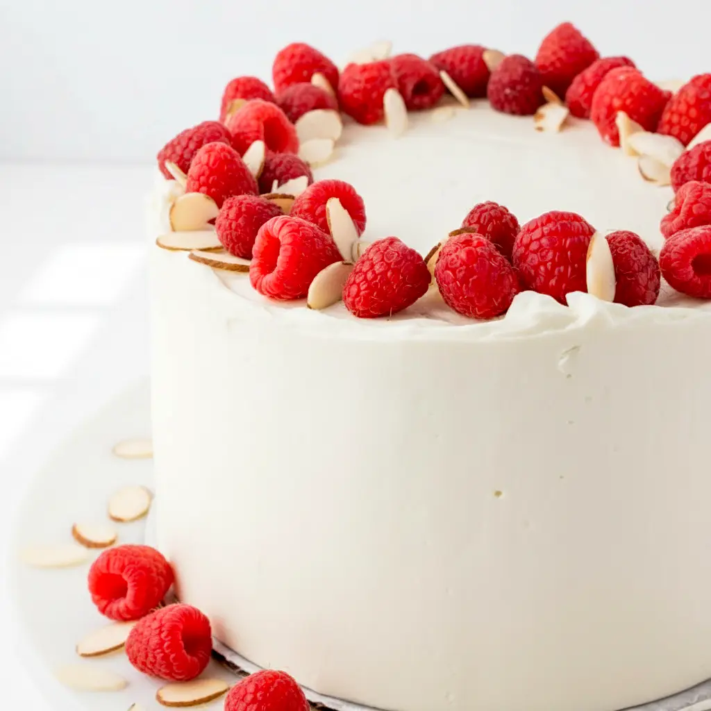 Raspberry Almond Cake with white almond cake layers, raspberry filling, creamy frosting, fresh raspberries, and slivered almonds on top.